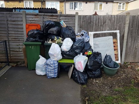 A collection of black and white rubbish bags, some with visible markings or tied at the top, are piled on and around a green bench situated on a paved area adjacent to a wooden fence, with an orange recycling bin and a green wheelie bin positioned behind the waste. To the right, there is a folded white frame or panel leaning against the fence, and a large green plastic container or basin is placed on the ground nearby. The scene appears to be part of a garden or back yard in Maida Vale, with a residential building partially visible in the background. The rubbish bags contain various forms of waste, and the overall setup suggests a garden clearance or rubbish removal effort by Garden Clearance Maida Vale, with the waste prepared for collection or disposal in accordance with local conditions in the area postcode.