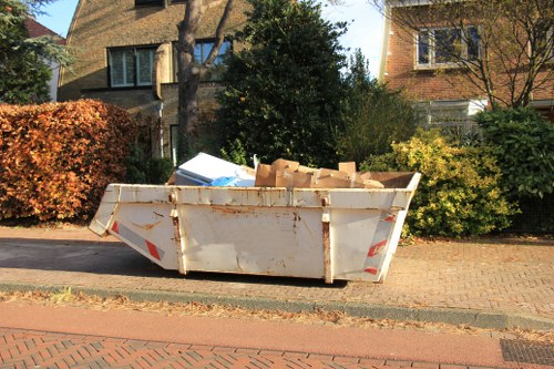 A large, white metal skip filled with a variety of cardboard boxes and waste materials, positioned on a paved driveway adjacent to a residential street. The skip shows signs of rust and weathering on its exterior, with the cardboard boxes stacked loosely inside, some overflowing slightly over the top edge. Surrounding the skip are suburban houses with brick facades, large windows, and sloped roofs, set amidst trees and bushes with autumn foliage, including a prominent dark green bush and a hedge with reddish-brown leaves. The scene is illuminated by natural daylight, casting soft shadows on the pavement and highlighting the textures of the cardboard and weathered skip, indicating routine rubbish removal within a residential area, such as Maida Vale or nearby in London, managed by Garden Clearance Maida Vale.