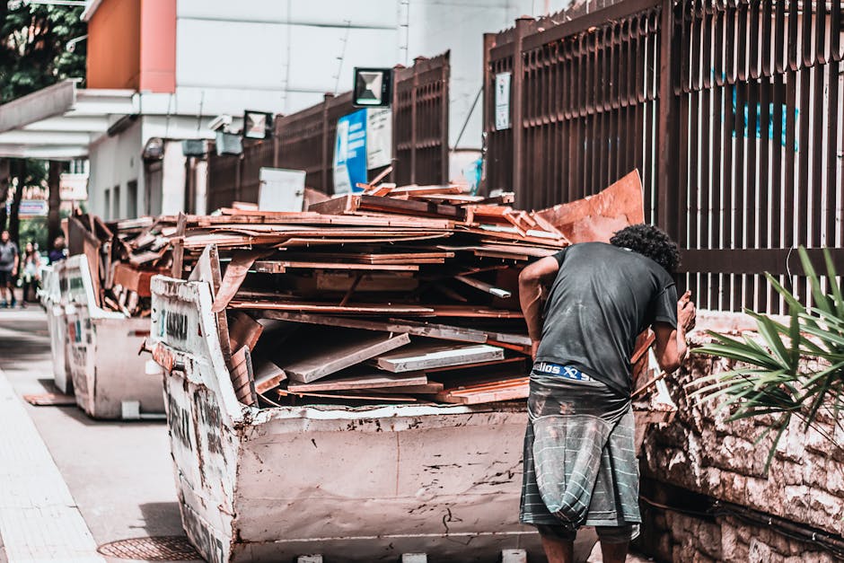 A small, dark red flatbed utility vehicle parked on a city street in front of a modern, curved glass-fronted office building. The truck's cargo bed is loaded with various bags of waste materials, incl