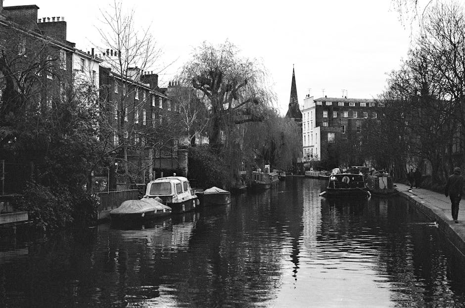A large, dark green boat is moored along the edge of a narrow canal, with its bow facing the viewer and showing signs of weathered varnish and surface grime. Behind it, there are two smaller boats, on
