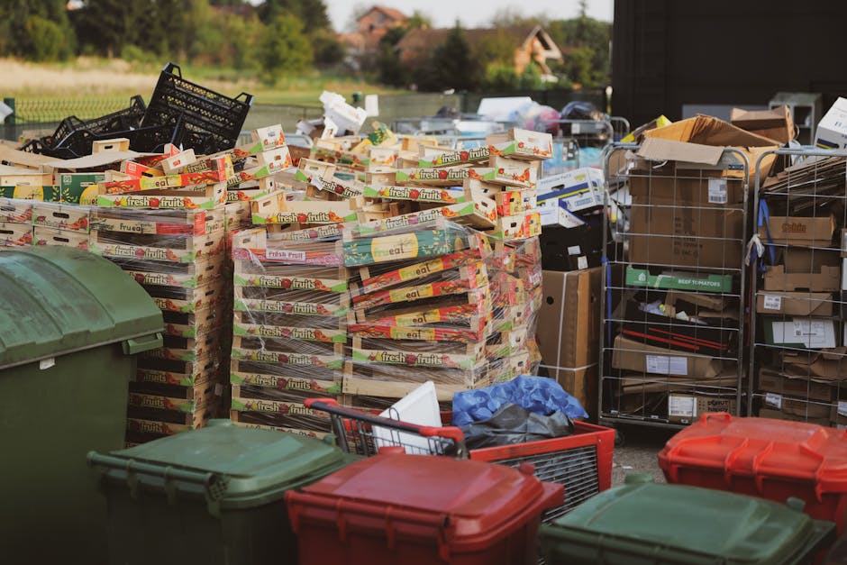 A collection of large, neatly stacked cardboard boxes filled with fresh fruit packaging, predominantly in red, green, and yellow tones, positioned outdoors on a paved surface. To the left, there are t