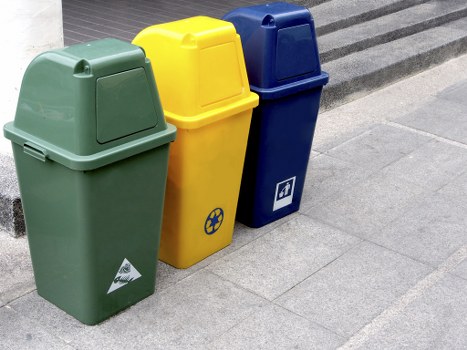 The image shows three wheeled rubbish bins positioned outdoors on a paved surface, likely on a street or driveway in a residential or commercial area. The first bin, on the left, is green with a textured finish, commonly used for general waste or recyclables and features a triangular recycling symbol on the front near the base. The middle bin is yellow with a smooth surface, typically designated for recycling specific materials, and has a circular icon with a recycling symbol on its front. The third bin, on the right, is dark blue with a similar smooth finish, often used for recyclable materials, and displays a white symbol resembling a document or paper on the front. All three bins are topped with closed lids, and they are aligned side-by-side, facing forward, with the background showing a curb or sidewalk edge. The setting suggests a typical collection point for rubbish removal services, such as those provided by Garden Clearance Maidavale, supporting local waste management in the area near Maidavale.