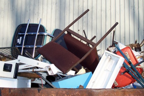 A large pile of assorted discarded items including a brown fabric office chair with wooden arms, a black plastic chair frame, a white metal-framed mirror, a blue plastic storage bin, and various pieces of red and blue plastic furniture, all stacked disorganizedly in an outdoor area against a light-colored corrugated metal wall. The scene appears to be part of rubbish removal or garden clearance efforts, with objects resting on a surface that suggests a driveway or yard, indicative of waste collection services such as those offered by Garden Clearance Maidavale in the local area postcode. The overall environment is cluttered, with objects varying in material, texture, and color, reflecting typical household and garden waste awaiting disposal.
